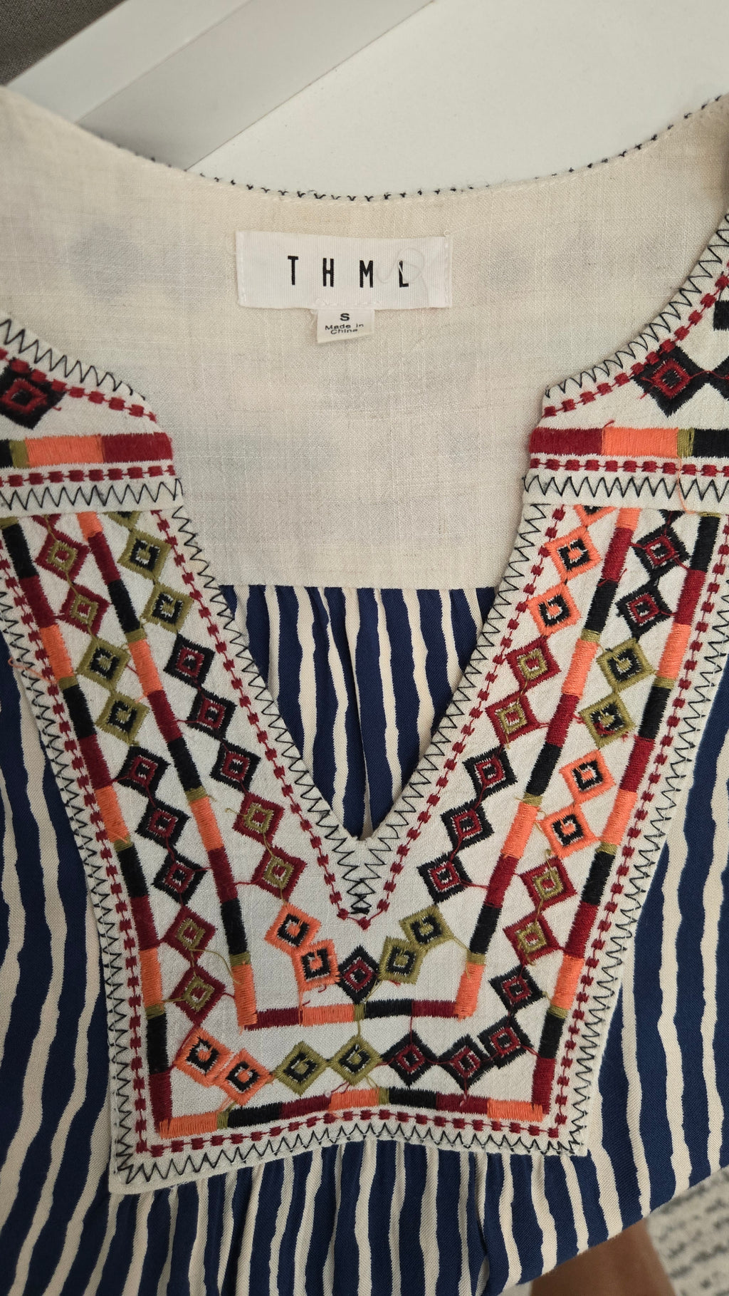 A close-up of a women's top with a V-neckline, featuring a cream-colored upper section and a navy blue and white vertically striped lower section. The front is decorated with a large, colorful geometric embroidered pattern in orange, red, black, and green.