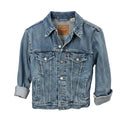 A light blue denim jacket displayed on a white hanger stand against a beige fabric backdrop. The jacket has a classic collar, button front, chest flap pockets, and rolled-up sleeves.